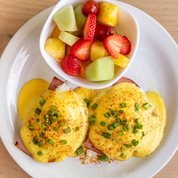 Cornerstone Cafe & Bar's Eggs Benedict with fruit salad, featuring hollandaise, ham, and chives.