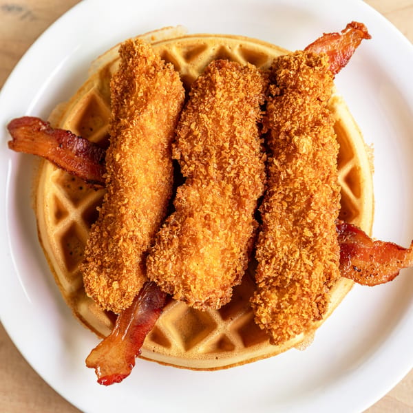 Golden-brown chicken and waffles with crispy bacon strips on a white plate at Cornerstone Cafe & Bar.