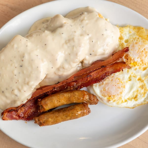 A breakfast plate with biscuits and gravy, crispy bacon, sausage links, and two fried eggs.