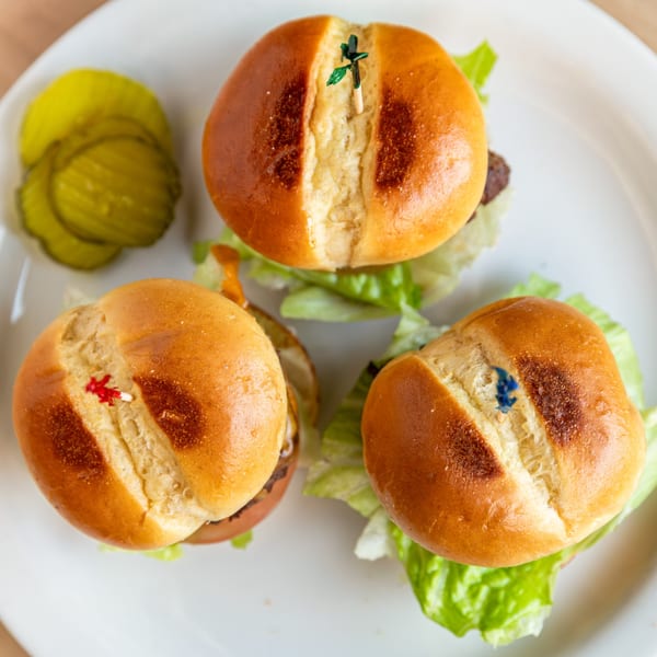 Three mini burgers with lettuce and pickles on a white plate, viewed from above.