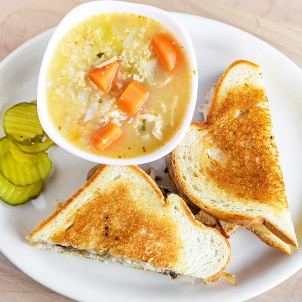 A bowl of chicken noodle soup with carrots, a grilled sandwich, and sliced pickles on a white plate.