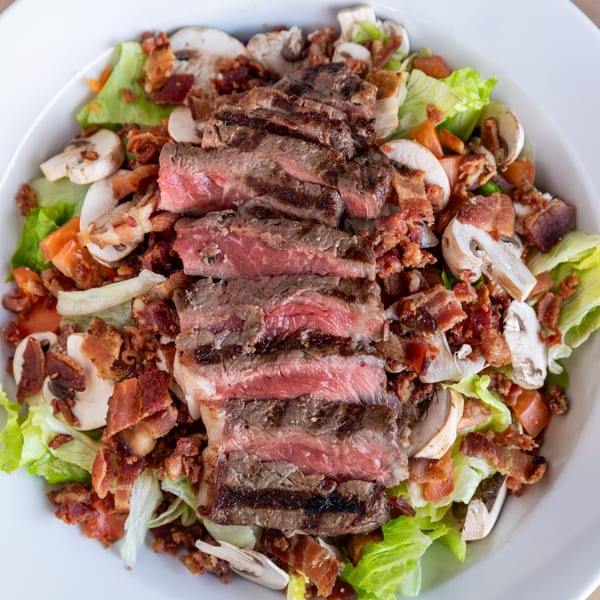 A close-up of a steak salad with sliced grilled steak, bacon bits, mushrooms, and lettuce.